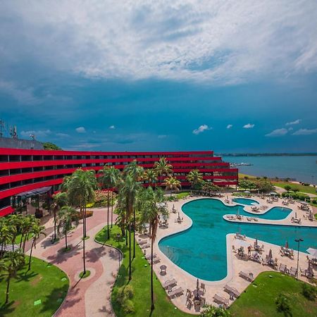 ホテル ロイヤル チューリップ ブラジリア アルボラーダ エクステリア 写真 The photo shows a modern hotel building with a striking red exterior, surrounded by lush greenery. In the foreground, there is a large, creatively shaped swimming pool, with lounge chairs arranged around it. The backdrop features a body of water, pos