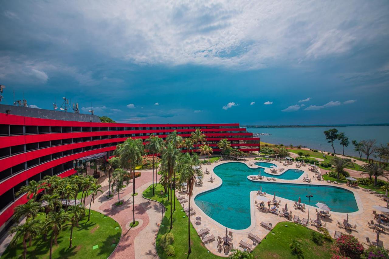 ホテル ロイヤル チューリップ ブラジリア アルボラーダ エクステリア 写真 The photo shows a modern hotel building with a striking red exterior, surrounded by lush greenery. In the foreground, there is a large, creatively shaped swimming pool, with lounge chairs arranged around it. The backdrop features a body of water, pos