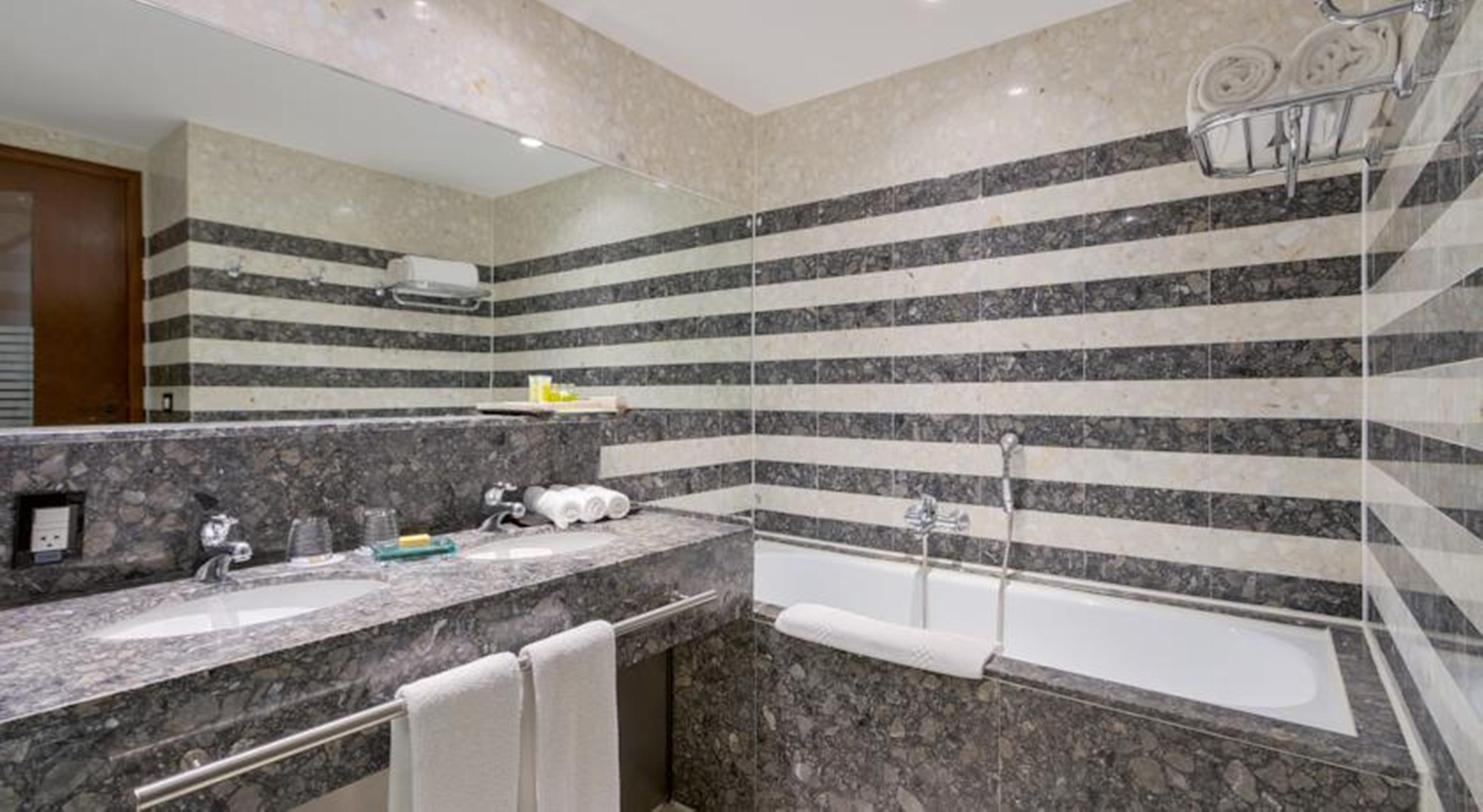 ホテル ロイヤル チューリップ ブラジリア アルボラーダ エクステリア 写真 The photo shows a modern bathroom featuring a combination of marble and stone finishes. The walls are decorated with horizontal stripes in various shades of gray and cream. There is a bathtub with a white exterior, and a showerhead is mounted on the 