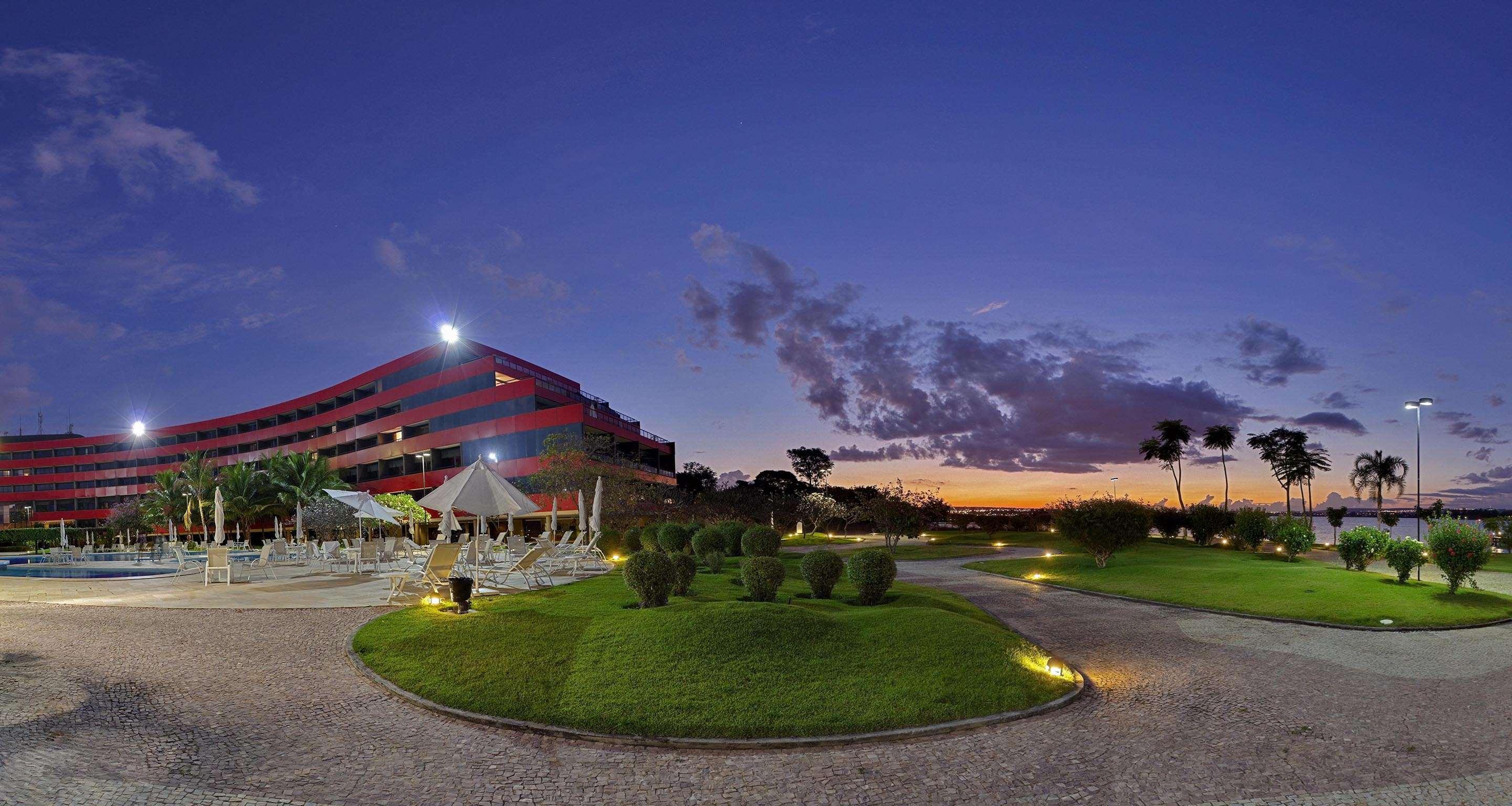 ホテル ロイヤル チューリップ ブラジリア アルボラーダ エクステリア 写真 The photo depicts a modern building, possibly a hotel or resort, with a striking red exterior. It is set in a landscaped area that features well-maintained greenery and ornamental plants. In the foreground, there are lounge areas with umbrellas, sugg