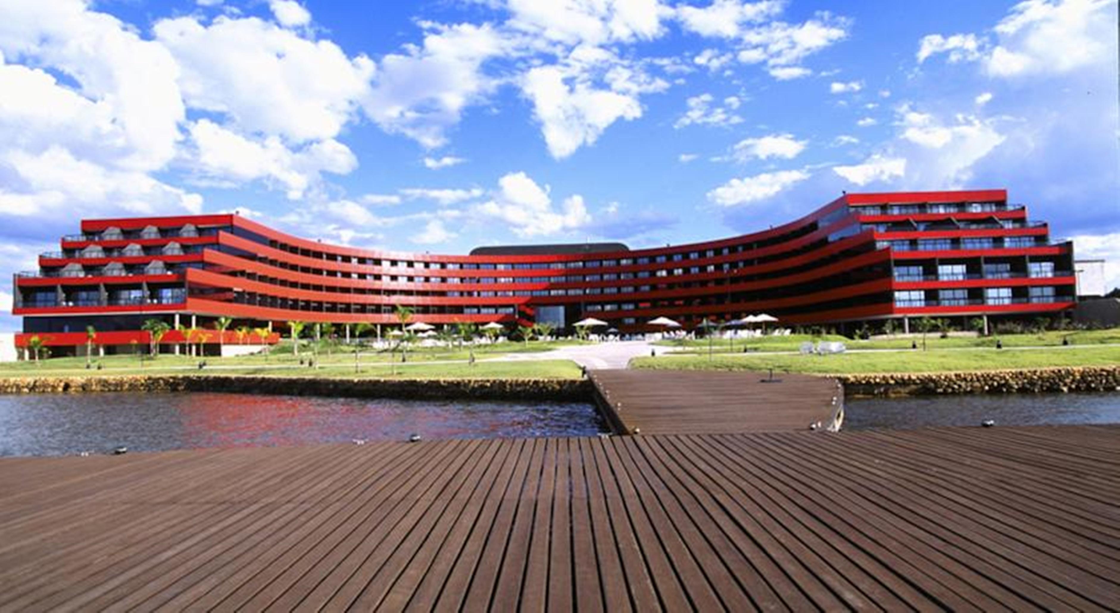 ホテル ロイヤル チューリップ ブラジリア アルボラーダ エクステリア 写真 The image shows a modern, curved building with a striking red facade, situated near a body of water. The architecture features multiple levels with large windows that create a sleek and contemporary appearance. In front of the building, there is a wo