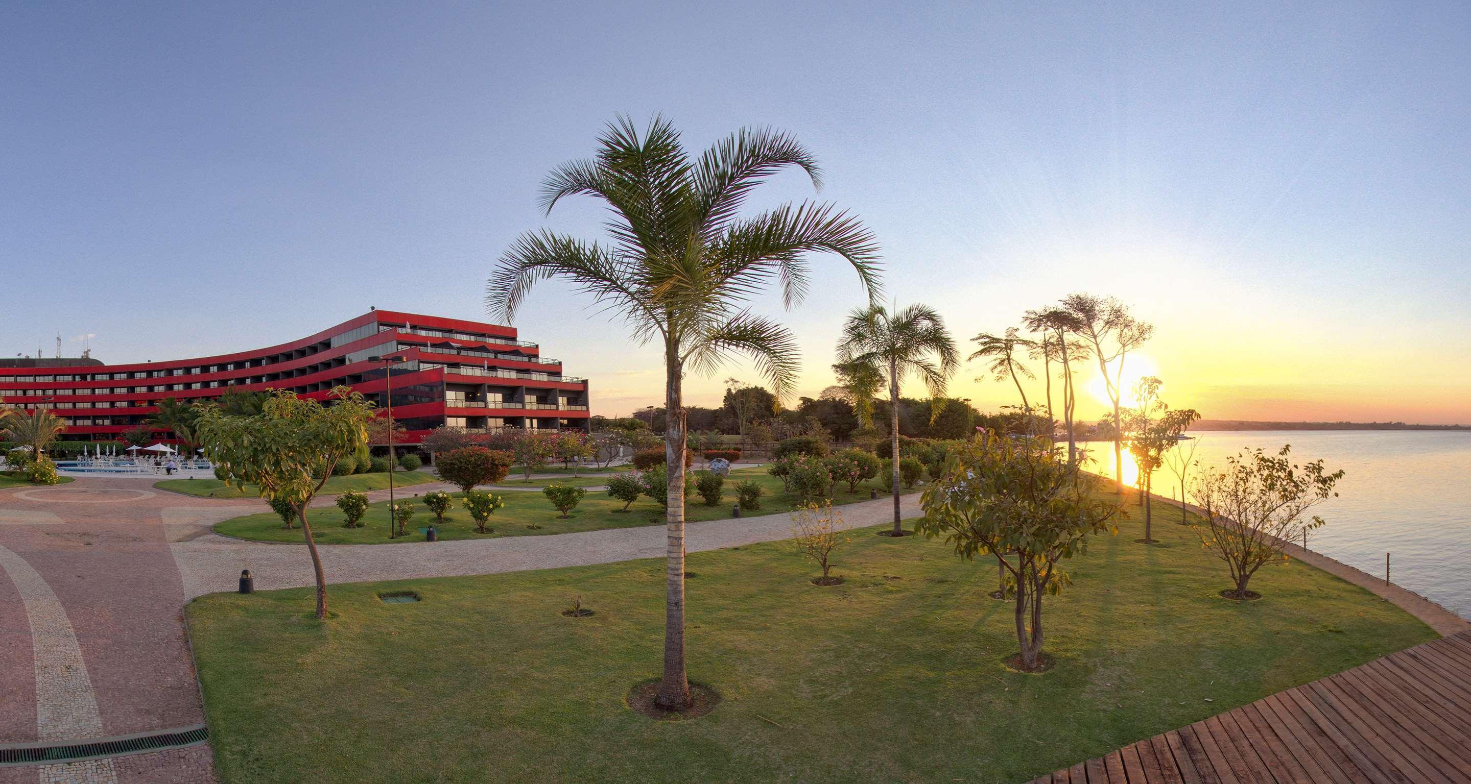ホテル ロイヤル チューリップ ブラジリア アルボラーダ エクステリア 写真 The photo depicts a scenic view featuring a red multi-story building alongside a serene waterfront. In the foreground, there are several palm trees and lush green lawns, creating a tropical atmosphere. The sun is setting in the background, casting wa