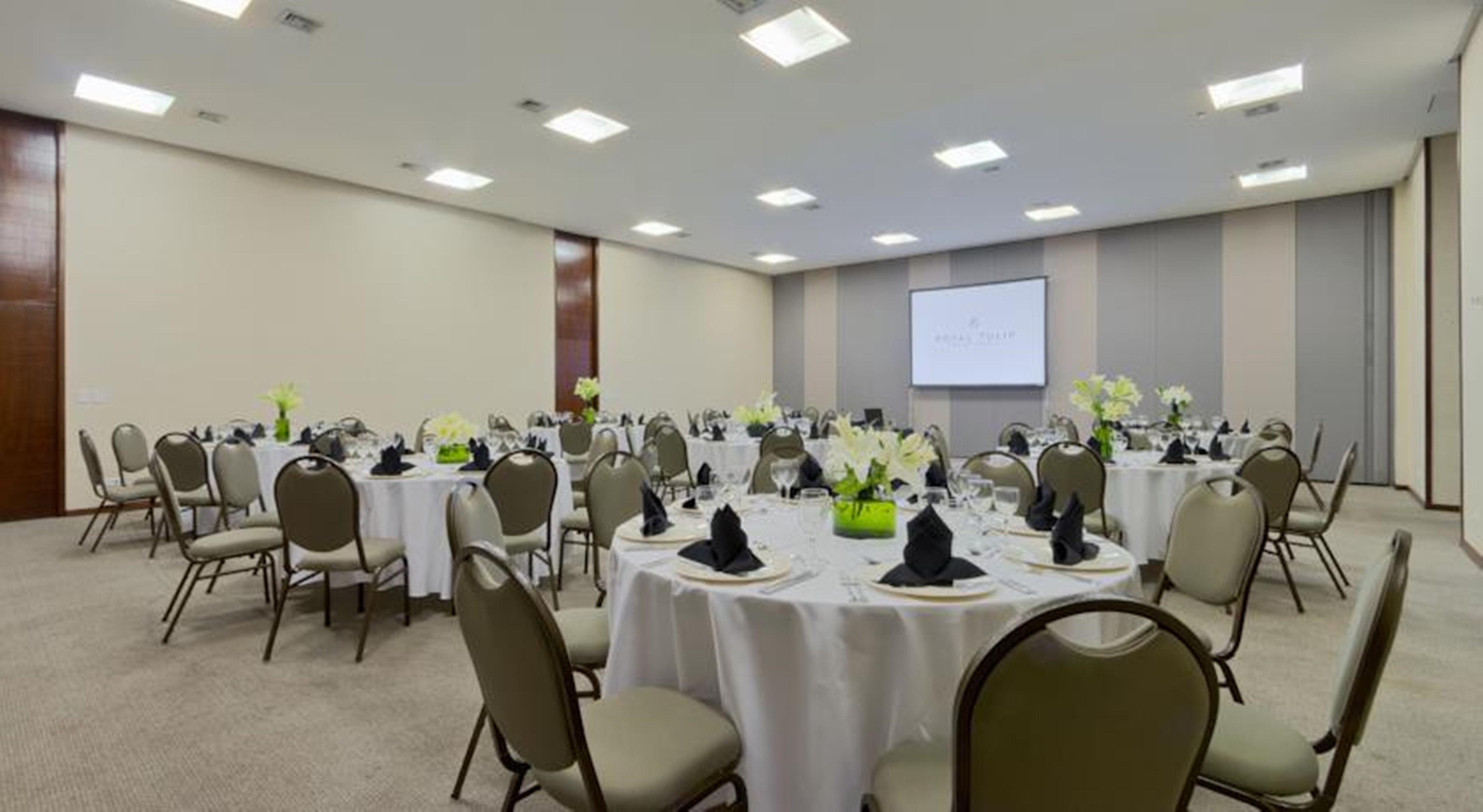 ホテル ロイヤル チューリップ ブラジリア アルボラーダ エクステリア 写真 The photo shows a banquet or conference room set up with multiple round tables covered in white tablecloths. Each table is arranged with black napkins and centerpieces featuring flowers, likely in shades of green and white. The room has beige walls a