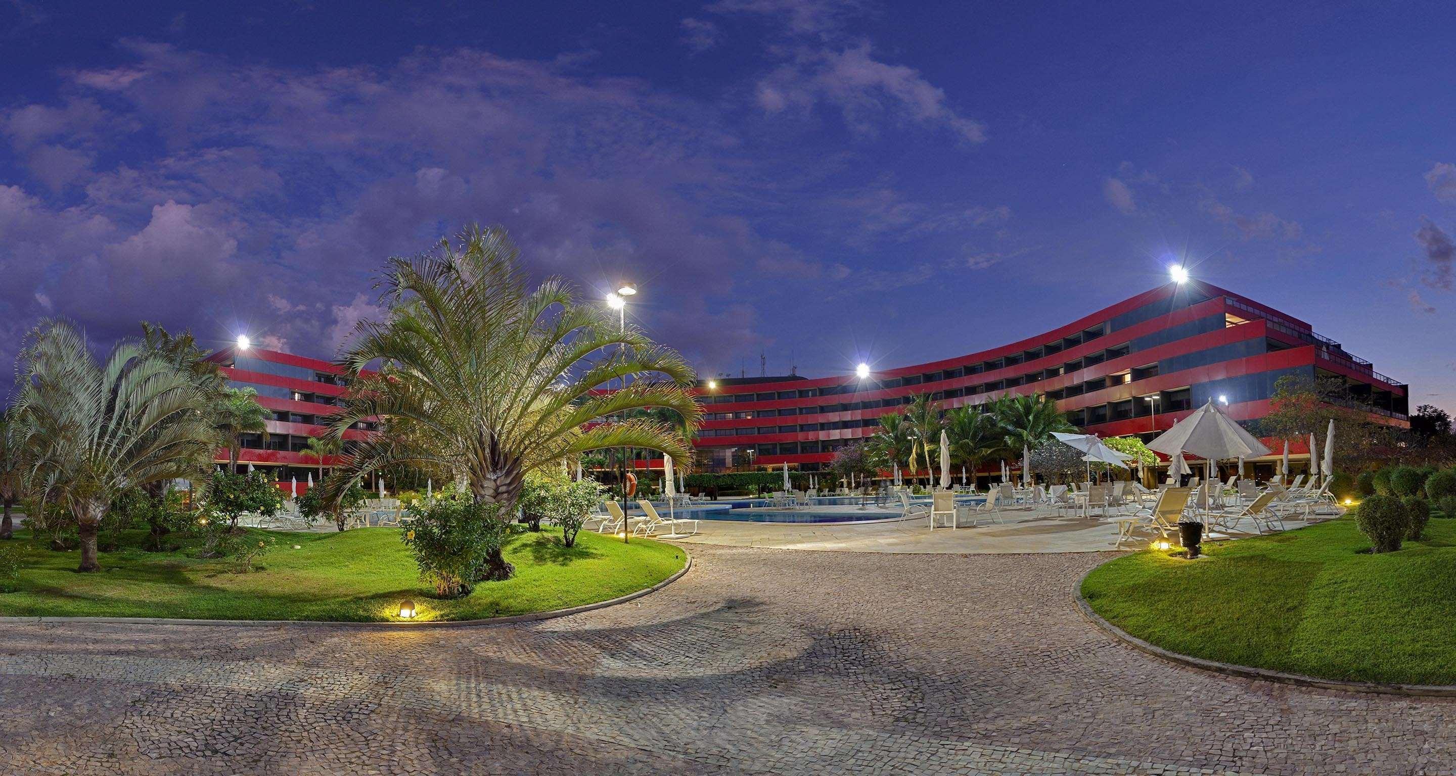 ホテル ロイヤル チューリップ ブラジリア アルボラーダ エクステリア 写真 The photo shows a panoramic view of a hotel or resort complex, characterized by its modern architectural design with smooth, curved lines and a vibrant red color. It’s set in a landscaped area featuring palm trees and green grass. The surroundings in