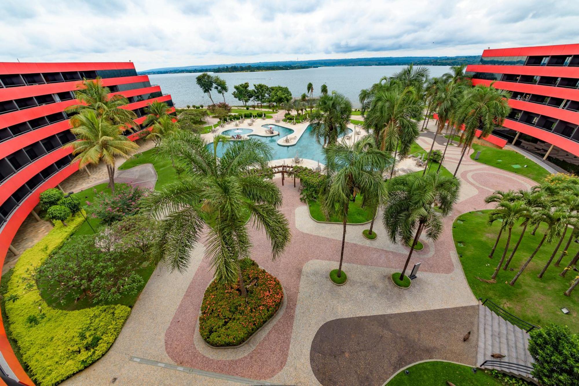ホテル ロイヤル チューリップ ブラジリア アルボラーダ エクステリア 写真 The photo shows a scenic view of a courtyard area in a residential complex. There are multiple red buildings surrounding a pool area, which is surrounded by palm trees and landscaped gardens. The pool features a curvy design, and there are pathways m