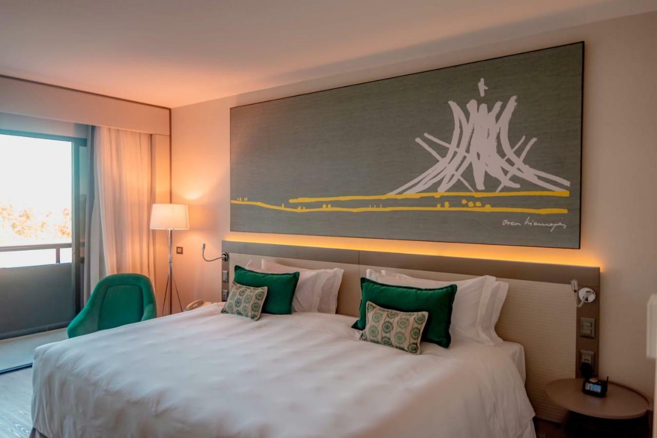 ホテル ロイヤル チューリップ ブラジリア アルボラーダ エクステリア 写真 The photo shows a modern hotel room featuring a large bed with white bedding and two green accent pillows. Above the bed, there is a large piece of artwork depicting a stylized mountain or volcanic shape with yellow accents. The room has a contempora